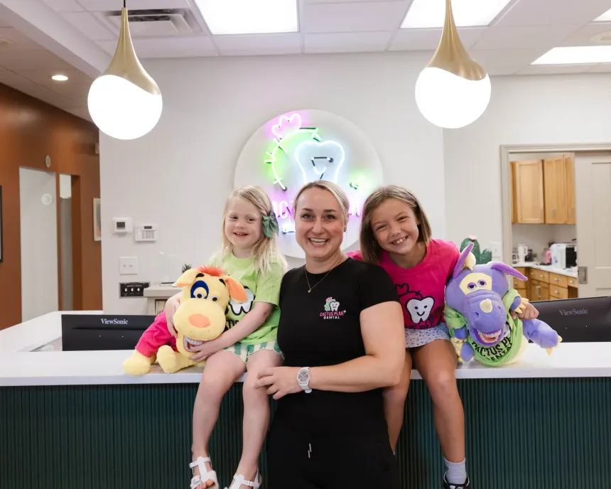 Dr. Kim Oberlander at the Cactus Pear Dental front desk with two young patients sitting on the counter beside her, holding the practice's cactus and dragon soft toys, the neon logo lit on the wall behind them