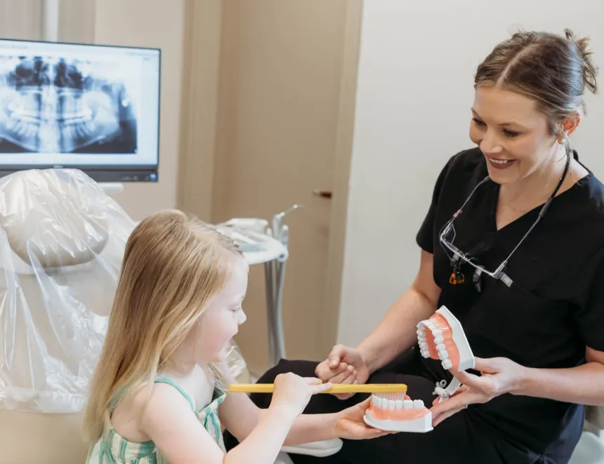 A young girl sitting up in the dental chair, holding an oversized toothbrush against a model of teeth that a smiling team member holds out for her